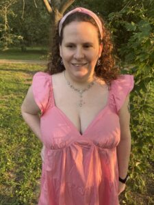 Daisy stands on grass near trees, with leafy branches filling most of one side of the image and early evening sunlight illuminating her, wearing an ankle-length bright pink dress with a deep v neckline, flutter sleeves, gathered fabric under the bust, and a tiered skirt, her curly auburn hair pulled back with a pink headband, and jewelry she can't describe because she doesn't remember what it is since this photo was taken months ago