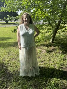 A woman with light skin and curly, light brown hair stands in the shade of a tree on a grassy lawn. She is wearing a sleeveless, light mint-green button-front top with a subtle texture, tied in a knot at the bottom. Over that, she has a long, sheer, pale green tulle skirt that lets the pattern of a shorter layer underneath show through. She has a light-colored headband and small hoop earrings, and there is a tattoo on her upper left arm. One hand rests on her hip and the other hangs relaxed at her side. Sunlight filters through the tree leaves above, creating dappled light and shadow on the grass around her, with more open lawn and a road in the background.